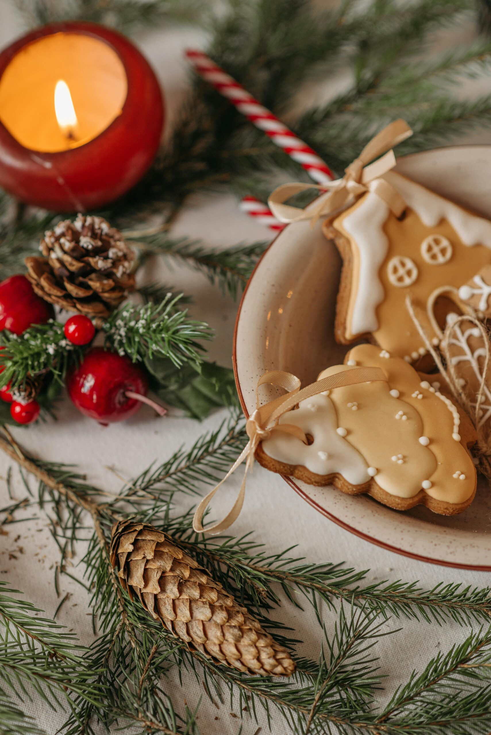 A festive table decorated with iced gingerbread cookies in a ceramic bowl surrounded by pine branches, pinecones, red berries, a lit red candle, and a striped candy cane.